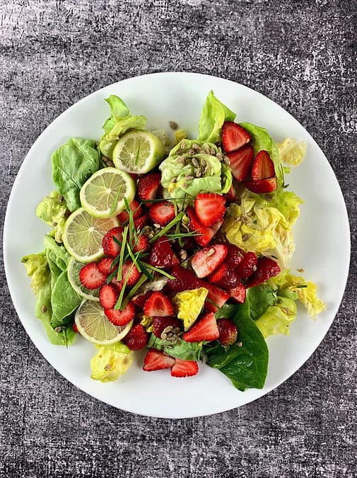 Lettuce salad with strawberries in a white salad platter with lime garnish in portrait view.