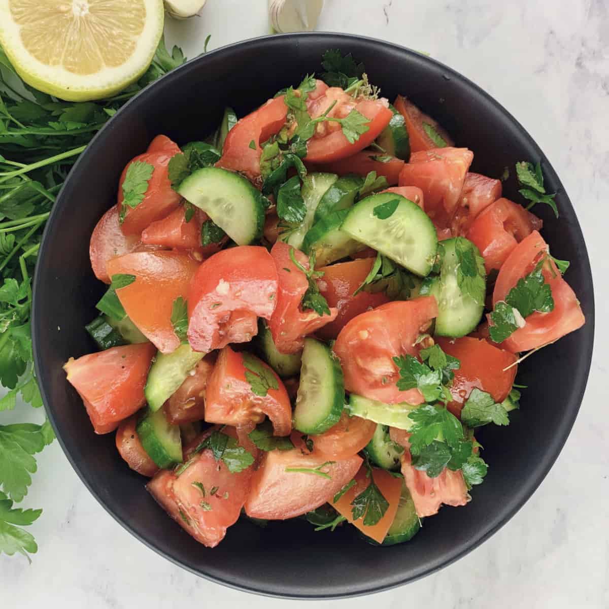 MEDITERRANEAN TOMATO CUCUMBER SALAD IN A BLACK BOWL WITH PARSLEY, LEMON AND GARLIC ON THE SIDE