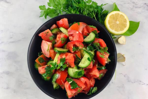 MEDITERRANEAN TOMATO CUCUMBER SALAD IN A BLACK BOWL WITH PARSLEY, LEMON AND GARLIC ON THE SIDE.