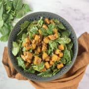 Pumpkin salad on a grey patterned shallow bowl with baby spinach in top left hand corner and orange linen napkin at the bottom.