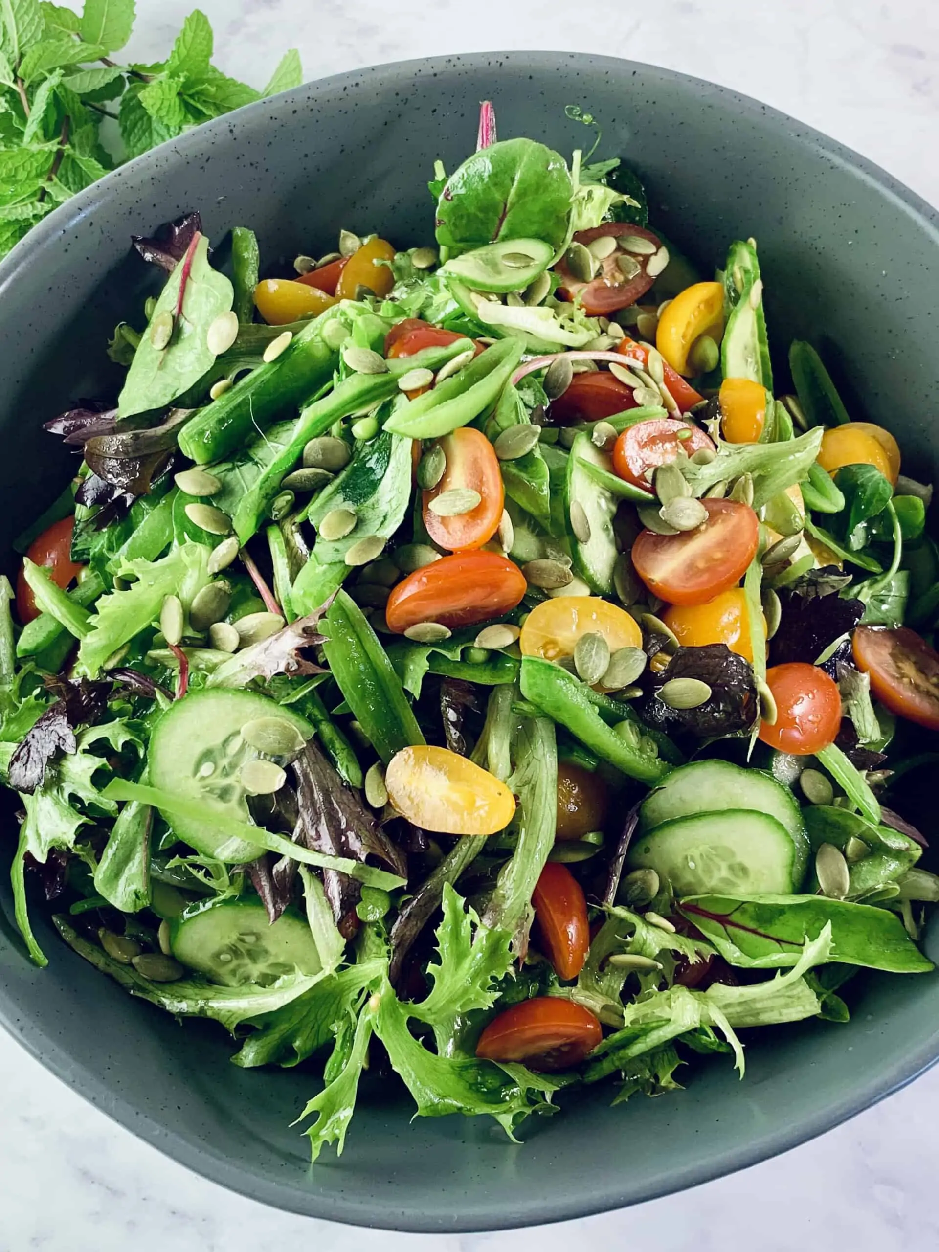 A close up of spring mix salad in a dark grey bowl with a sprig of mint in top left corner.