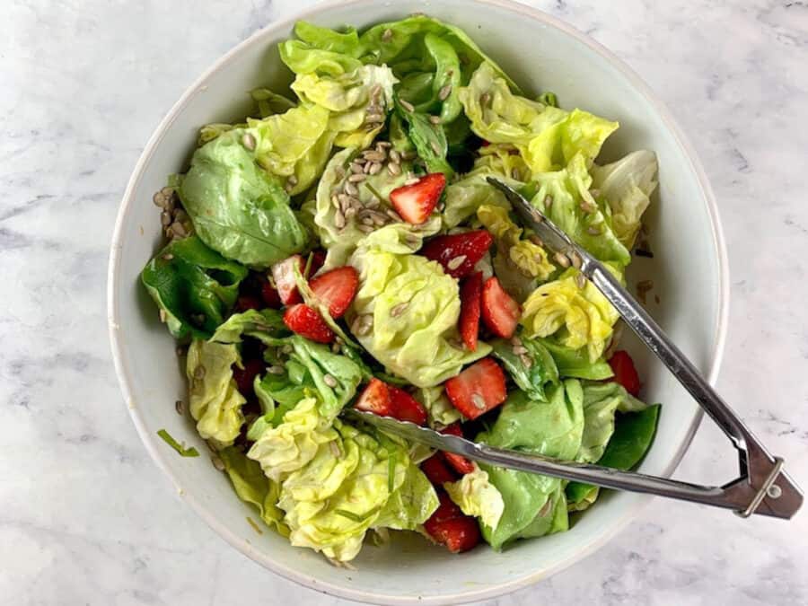 Tossing lettuce salad with strawberries in a white bowl with tongs.