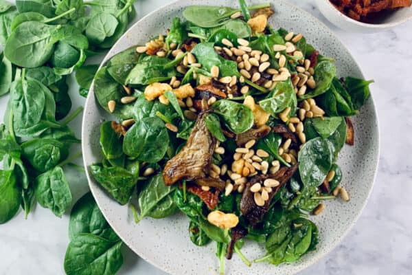 Oyster Mushroom Salad on a ceramic plate with spinach leaves scattered on the left and sundried tomatoes in a bowl on the top right.