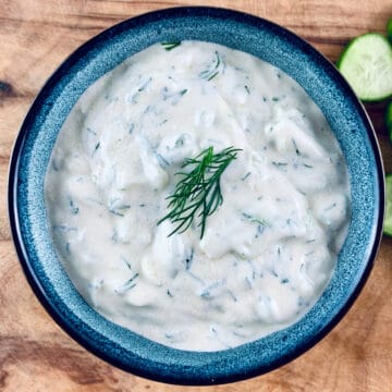 Our Authentic Tzatziki Recipe in a blue bowl with cucumber slices on top right sitting on a wooden board.