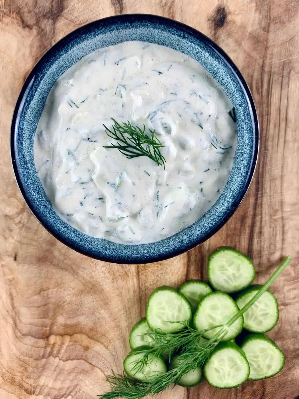 Our Authentic Tzatziki Recipe in a blue bowl with cucumber slices and dill sprig on the bottom left and sitting on a wooden board.