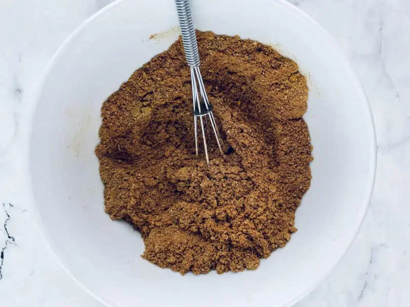 Mixing ras el hanout spices in a white bowl with a small whisk.