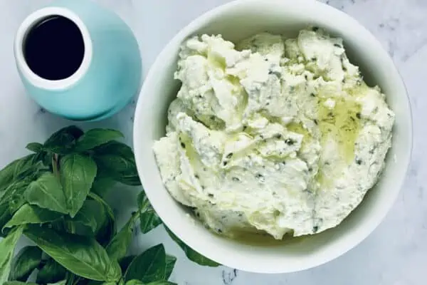 Whipped feta in a bowl with fresh basil and olive oil in an aqua bottle on left.