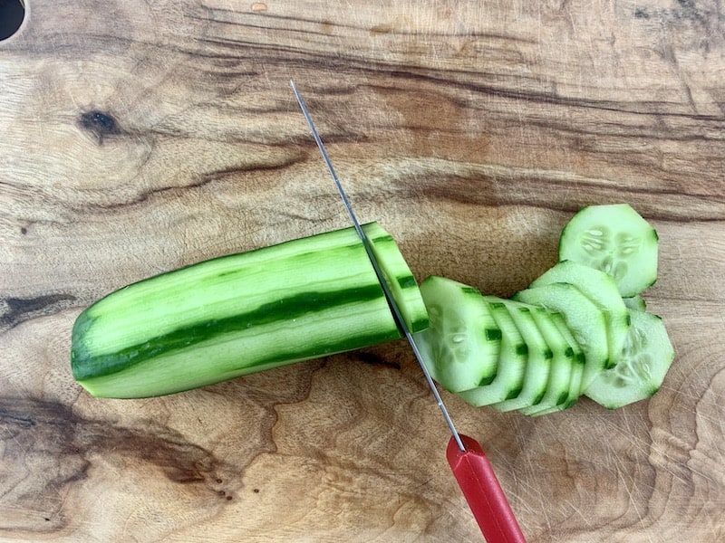 Peeled cucumber on wooden chopping board being sliced into rounds.