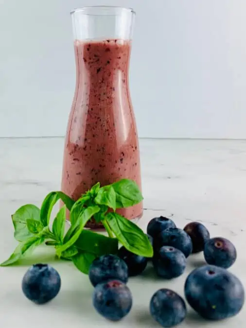 Blueberry salad dressing in a glass bottle with fresh blueberries and basil sprigs in front.