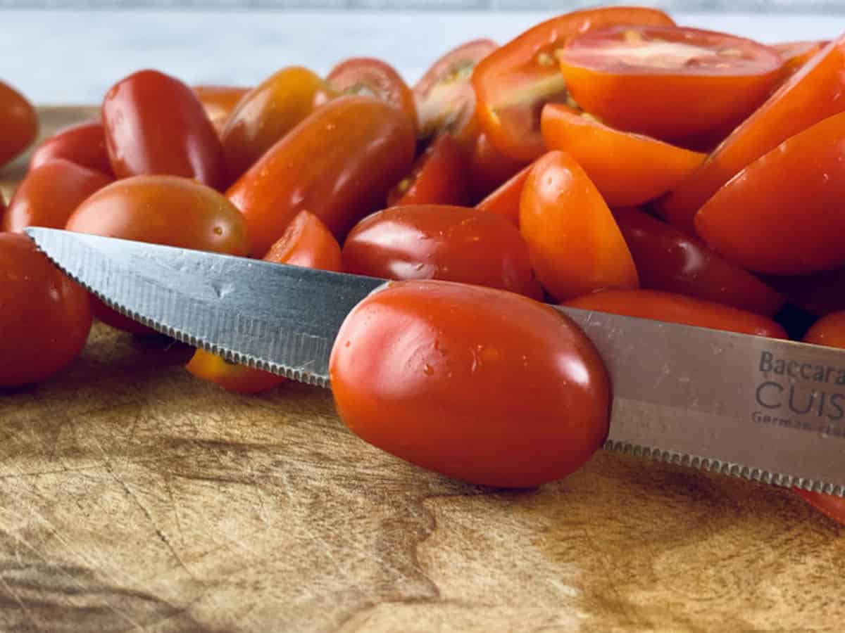 A close-up of a mini Roma tomatoes being cut with a knife on a wooden board.