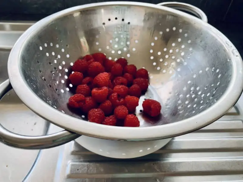 Raspberries draining on sink in a colander with a plate underneath.