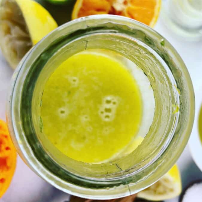 Shaken citrus salad dressing in glass jar with ingredients scattered around.