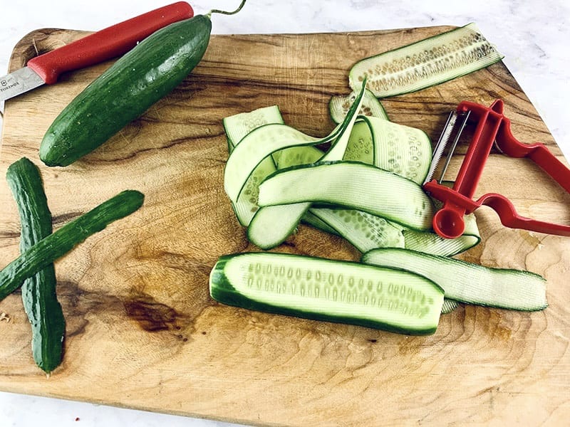 Cucumber being cut into ribbons on a wooden board with a vegetable peeler.