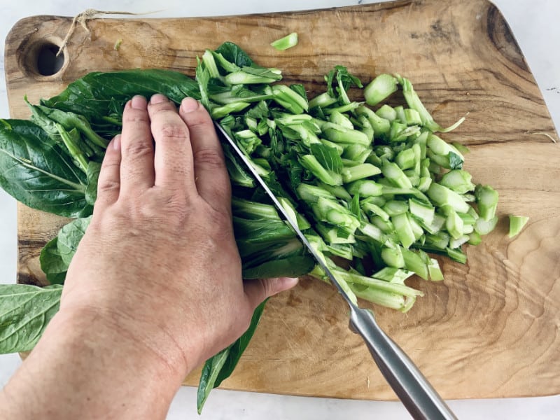 Hands cutting choy sum on a wooden board.