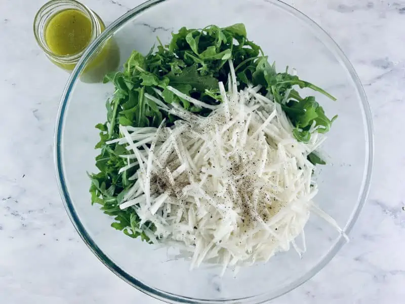 Grated Kohlrabi and rocket in a glass bowl with dressing at top left.