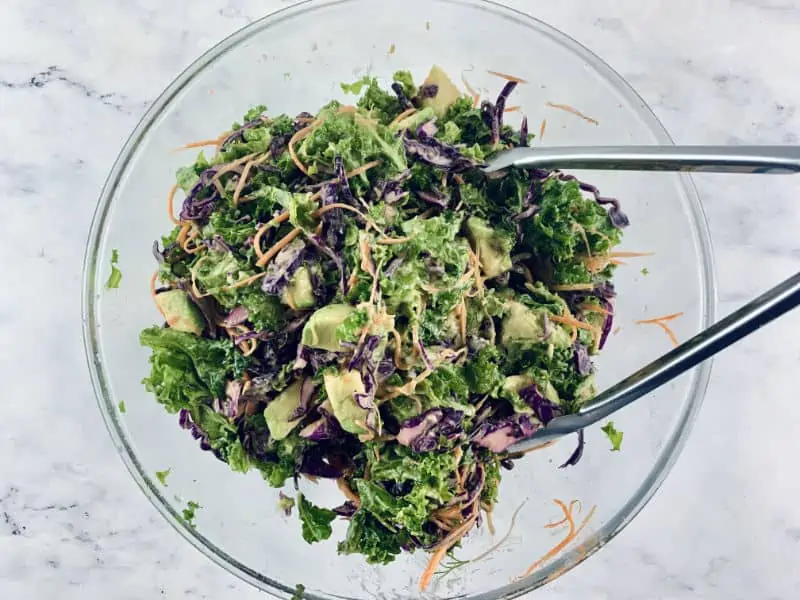 Tongs mixing kale coleslaw with avocado mayo in a glass bowl.