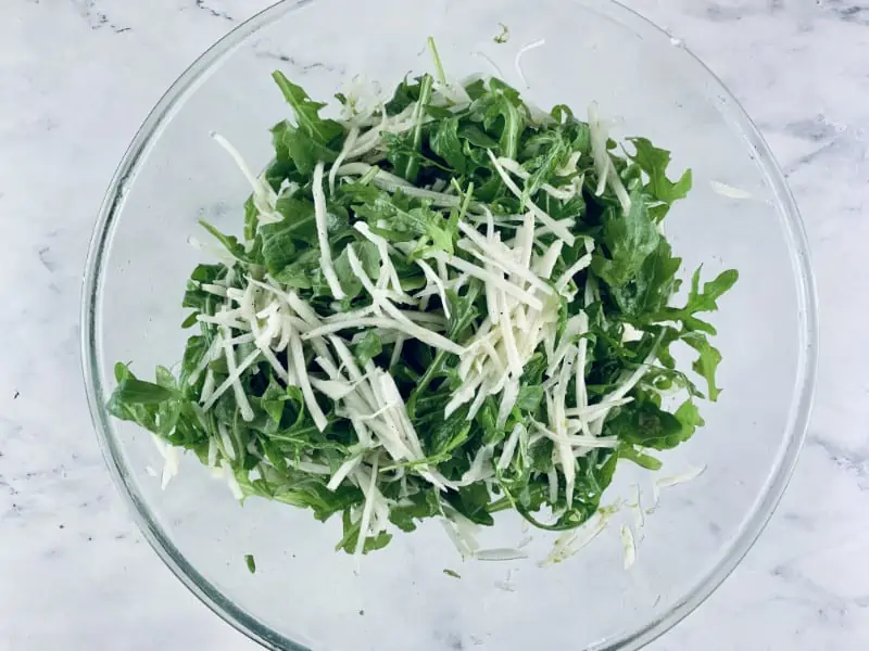 Tossed grated kohlrabi, rocket and lime dressing in a glass bowl.