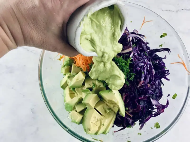 Hand pouring avocado mayo over kale coleslaw.