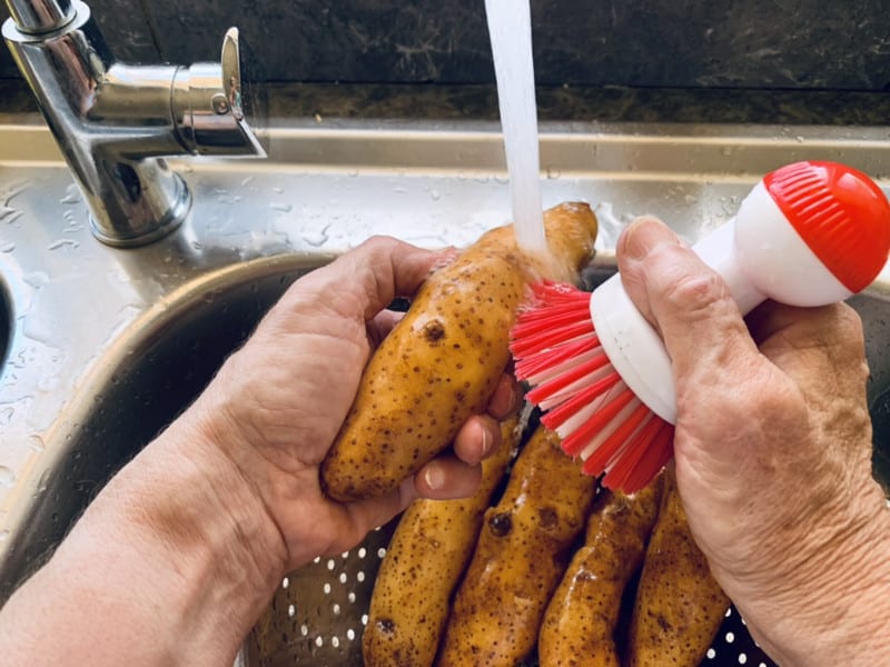 Hands scrubbing kipfler potatoes under cold water in a sink.
