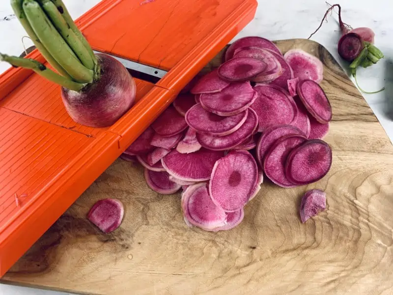 Watermelon radish being sliced with a mandoline slicer on wooden board.