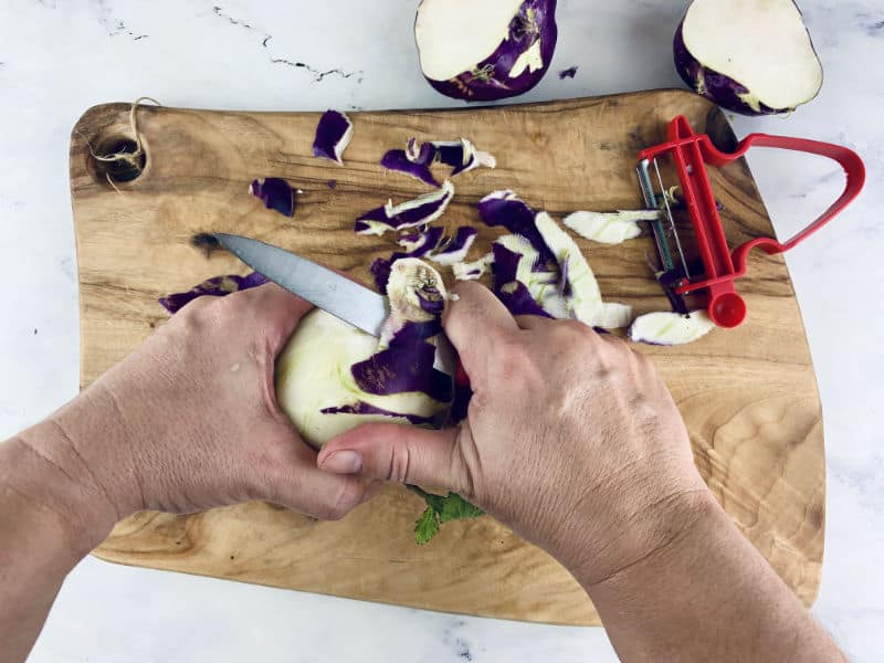 Hands trimming kohlrabi bulb on a wooden chopping board.
