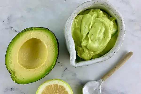 Avocado mayo in a white pottery jug with avocado half, lemon half and spoon on the side