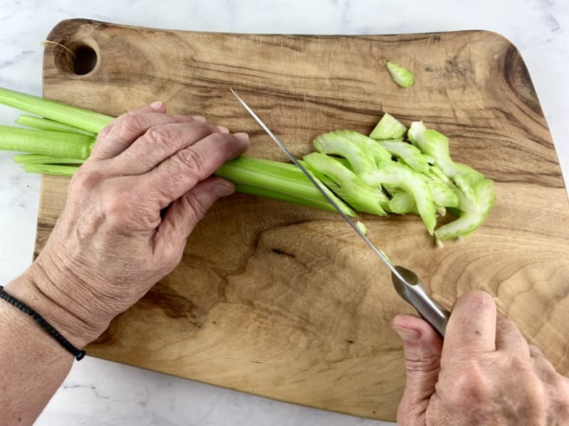 Hands slicing green celery stalks on a wooden board with a knife.