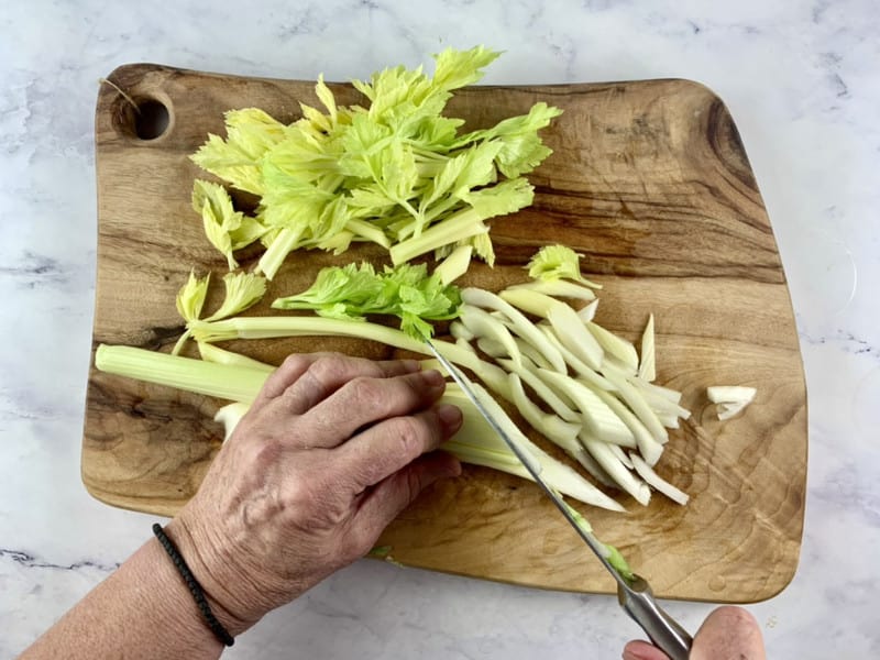 Hands slicing yellow celery stalks on wooden board with knife.