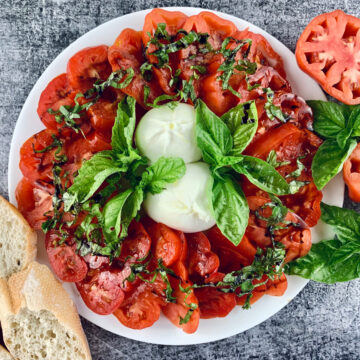 Burrata caprese salad with sliced tomatoes, bresh bread and basil sprigs on the side.