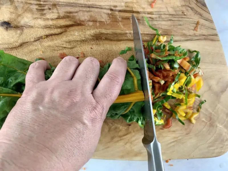 Rolled chard leaves being held and sliced thinly with a knife on a wooden board.