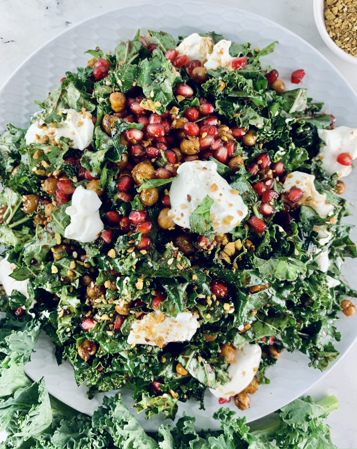 A close-up of Kale pomegranat salad on a white plate with kale and dukkah on the side.