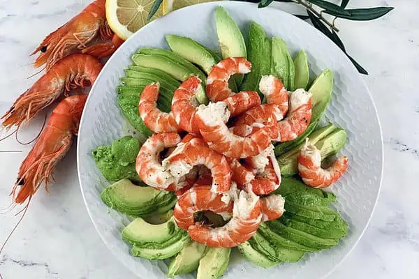 Prawn avocado salad on a white plate with prawns and lemon slices on the side.