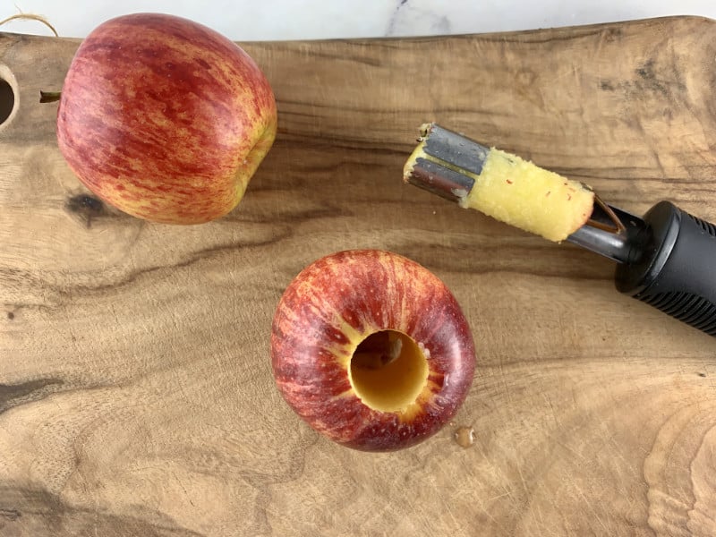 Cored apple with apple corer on a wooden board and a red apple on the side.