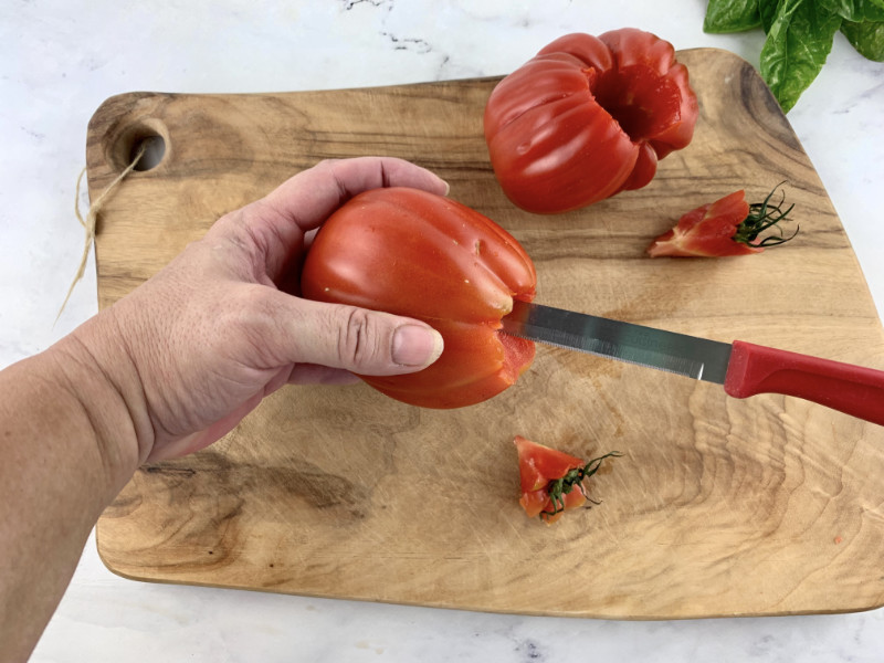 Hands removing tomato core with a knife on a wooden board.