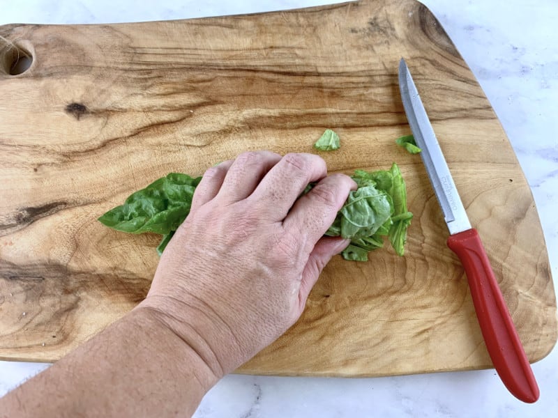 Hands rolling stak of basil leaves into a cigar shape on a wooden board with a knife on the side.