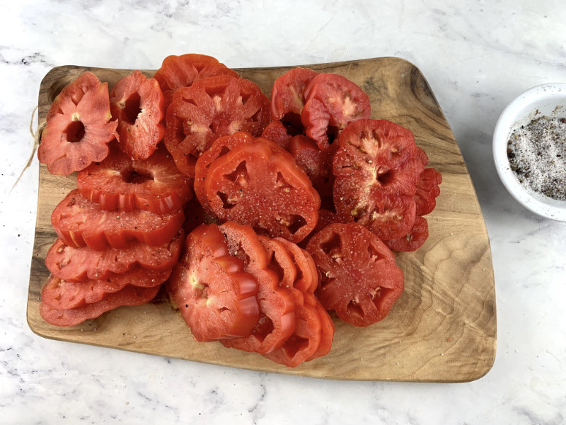 Salted heirloom tomatoes sitting on a wooden board with salt and pepper on the side.