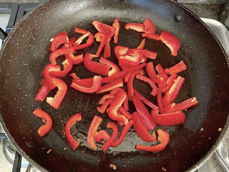 Red capsicum strips being sauteed in pan.