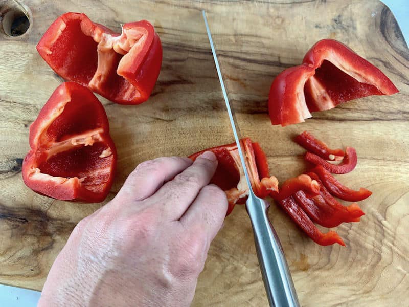 Hands slicing red capsicum into strips on a wooden board.