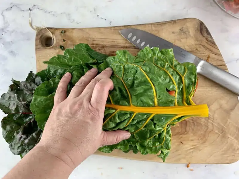 Hands stacking rainbow chard leaves on a wooden board, knife on the right.