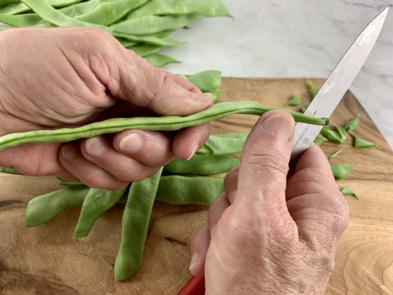 Hands stringing flat beans with a knife on a wooden board.