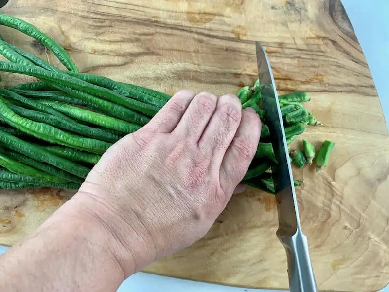 Hands trimming snake beans on a wooden board.