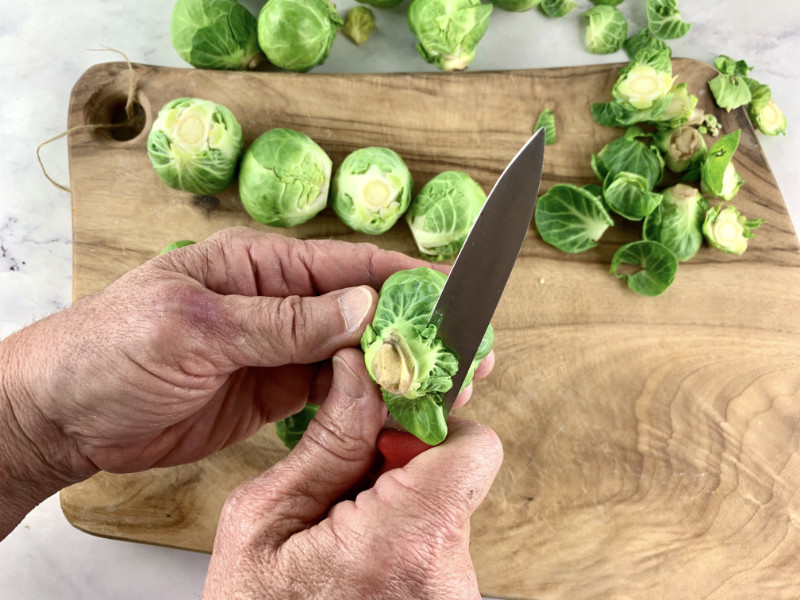 Hands trimming sprouts on a wooden board with a knife.