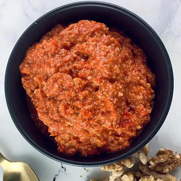 Our Lebanese Muhammara recipe in black bowl with gold spoon and walnuts on the side.
