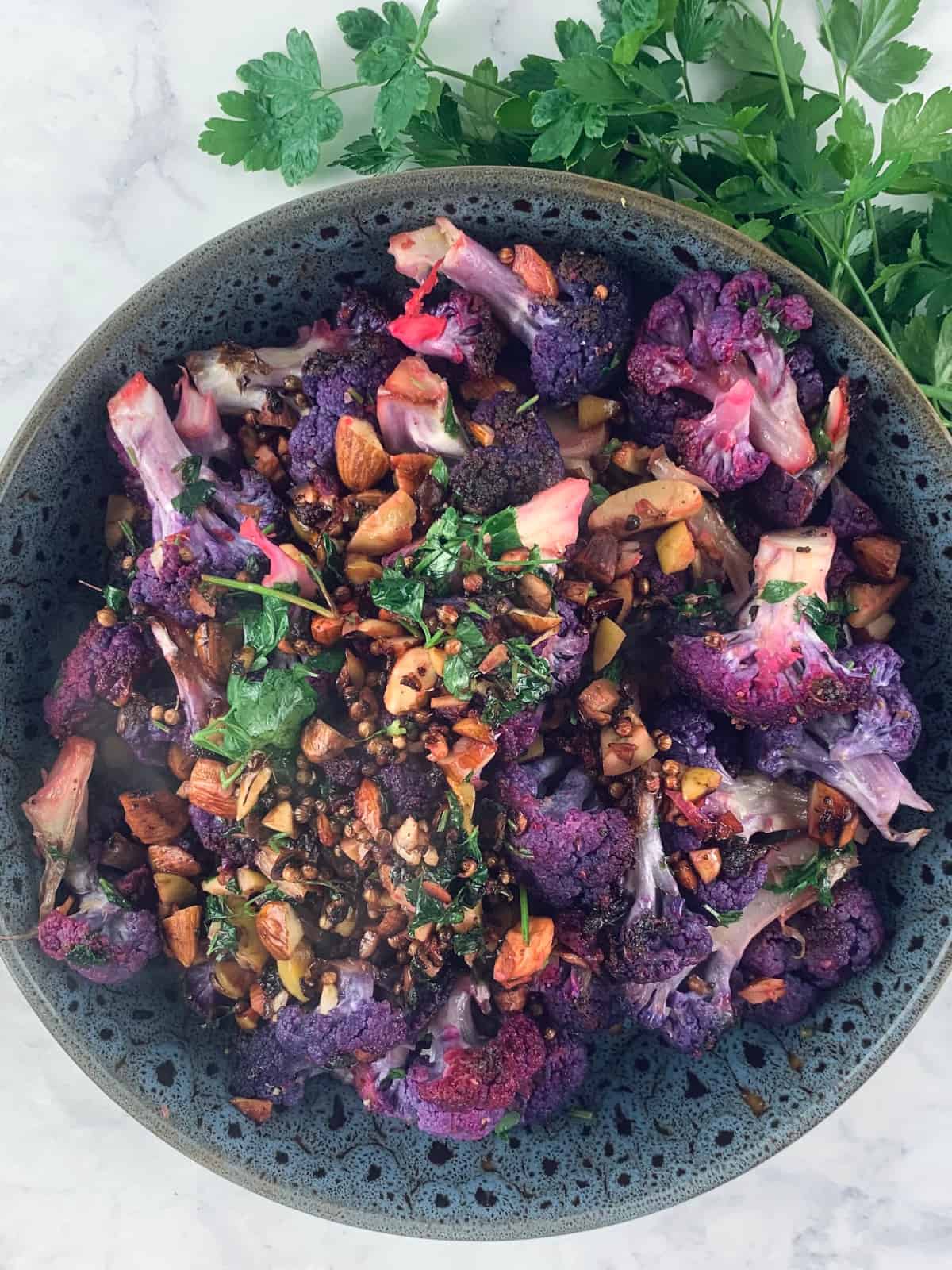 Purple cauliflower salad in shallow dark grey patterned salad bowl with parsley sprigs on the top right corner.
