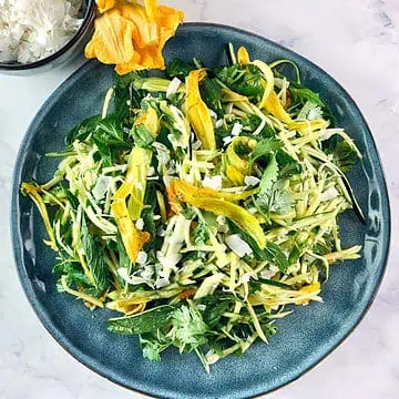 Asian zucchini salad on a blue plate with zucchini flowers and coconut flakes on the side.