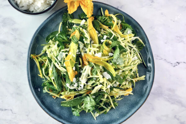 Asian zucchini salad on a blue plate with zucchini flowers and coconut flakes on the side.
