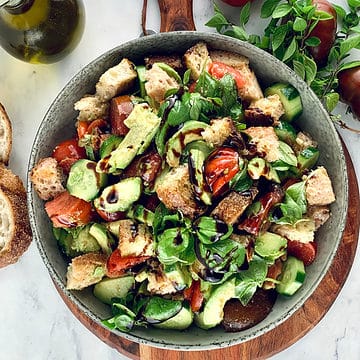 Avocado panzanella in a ceramic bowl on top of a wooden board, with ingredients around.