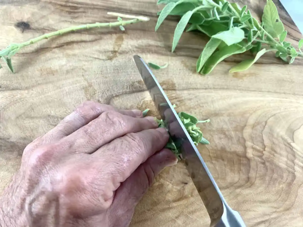 Hands chopping sage leaves with a knife on a wooden board.