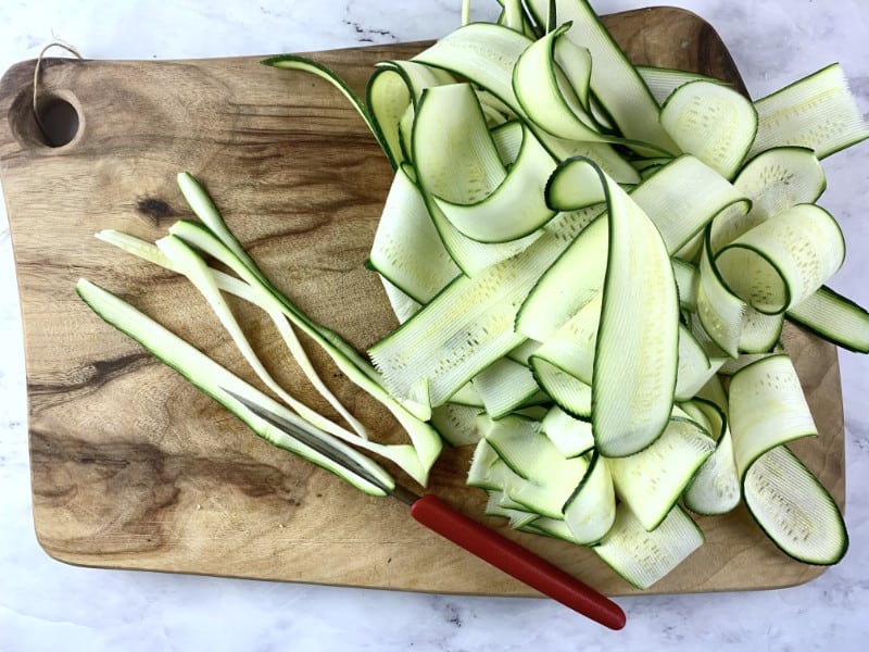 Cutting zucchini ribbons into thin spaghetti. on a wooden chopping board with a knife.