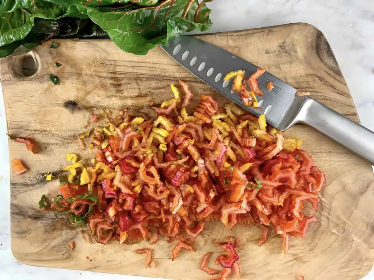 Finely chopped chard stems on a wooden board with a knife.